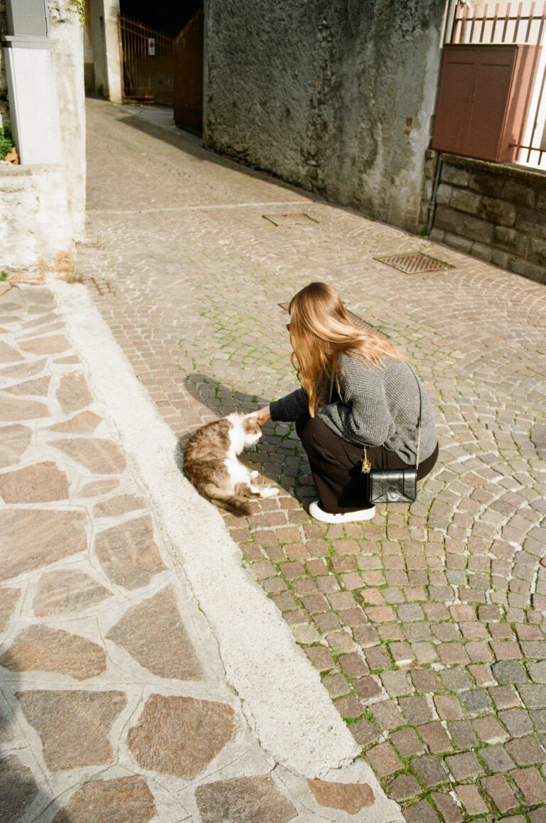 A woman petting a cat on a sunlit cobbled street